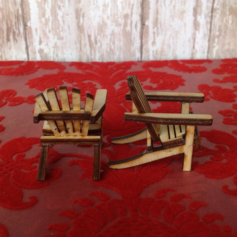 May include: Two miniature wooden chairs in a classic adirondack style, with a natural wood finish. The chairs are sitting on a red velvet surface.