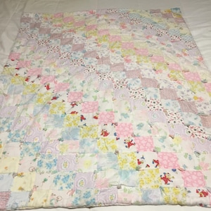 May include: A patchwork quilt with a variety of pastel-colored squares. The squares feature floral, polka dot, and cartoon character patterns in pink, blue, yellow, and lavender. The quilt is laid out on a white surface.