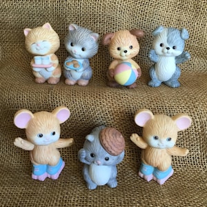 May include: A collection of seven ceramic animal figurines. The figurines include cats, dogs, and mice, each with unique features and accessories. The color palette includes shades of tan, gray, blue, and pink. The figurines are displayed on a textured, brown surface.
