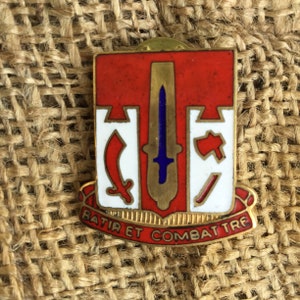 May include: A rectangular pin with a red and white design, featuring a sword, axe, and saber. The pin has gold trim and the words "BATIR ET COMBATTRE" in red on a gold banner.