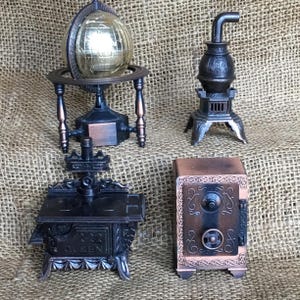 May include: Four miniature metal sculptures: a globe, a stove, a safe, and a potbelly stove. The globe is gold-colored, and the others are bronze and black. The safe has a combination dial and decorative scrollwork. The stove has the word "QUEEN" on it.