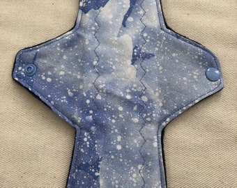 May include: A blue and white reusable cloth menstrual pad with a white splatter pattern. The pad has black edging and two blue snap closures.