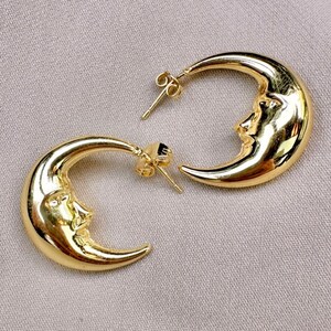 May include: A pair of gold-coloured crescent moon earrings. Each earring features a detailed face carved into the moon's surface. The earrings have a post-and-clutch closure. The background is a neutral, light tan colour.