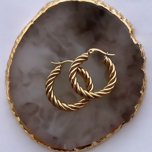 Puede incluir: Un par de pendientes de aro dorados con un diseño de cuerda trenzada. Los pendientes se muestran sobre una superficie de ágata blanca y dorada.