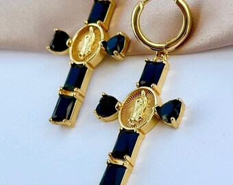 Puede incluir: Pendientes de aro dorados con un dije de cruz colgante. La cruz está adornada con piedras de circonita cúbica negras y un medallón dorado con una figura religiosa.