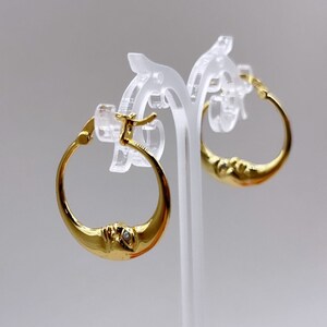 May include: Gold-colored hoop earrings with a crescent moon design. Each earring features a detailed face on the moon's curve and a small, clear gemstone. The earrings are displayed on a clear stand against a light background.