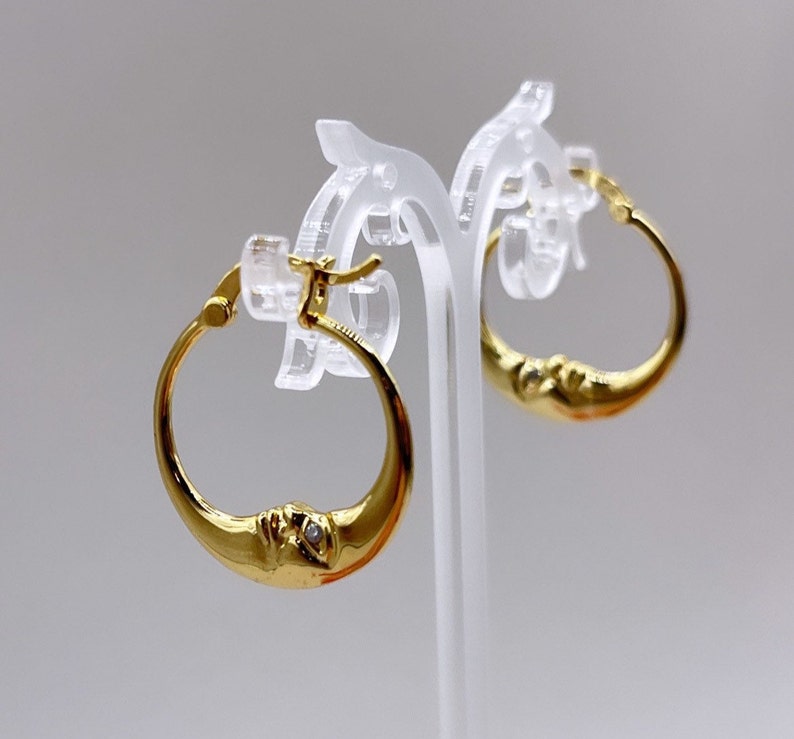 May include: Gold-colored hoop earrings with a crescent moon design. Each earring features a detailed face on the moon's curve and a small, clear gemstone. The earrings are displayed on a clear stand against a light background.