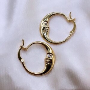 May include: Gold-colored hoop earrings with a crescent moon design. Each earring features a moon face with a small, clear gem. The text "MOON FACE DESIGN ON BOTH SIDES*" is visible at the top.