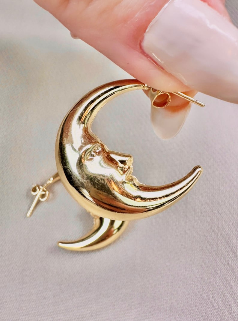May include: Gold-coloured crescent moon earrings. Each earring features a detailed face on the moon's surface. The earrings are held up against a light-coloured background, showcasing their shiny, reflective surface.