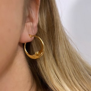 May include: Gold-colored crescent moon hoop earring with a small, clear gemstone. The earring features a detailed face design on the moon. The earring is worn on a person's ear, with blonde hair visible in the background.