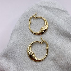 May include: A pair of gold-colored hoop earrings. Each earring features a crescent moon design with a face carved into the moon. A small, clear stone is set into the face. The earrings are displayed on a light gray surface.