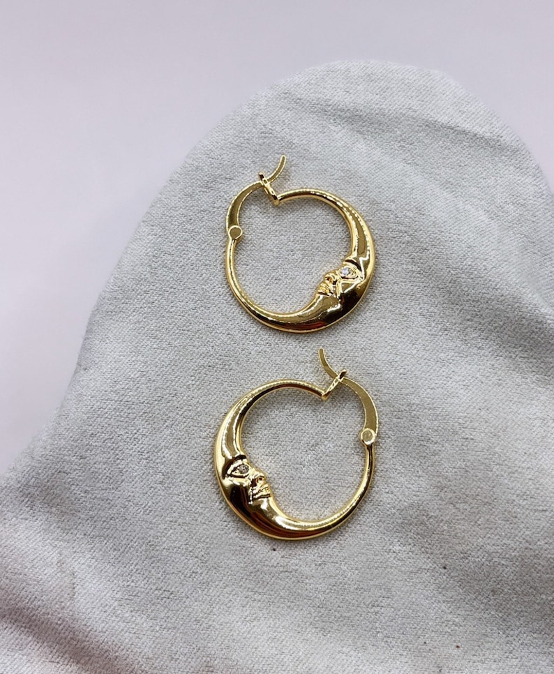 May include: A pair of gold-colored hoop earrings. Each earring features a crescent moon design with a face carved into the moon. A small, clear stone is set into the face. The earrings are displayed on a light gray surface.