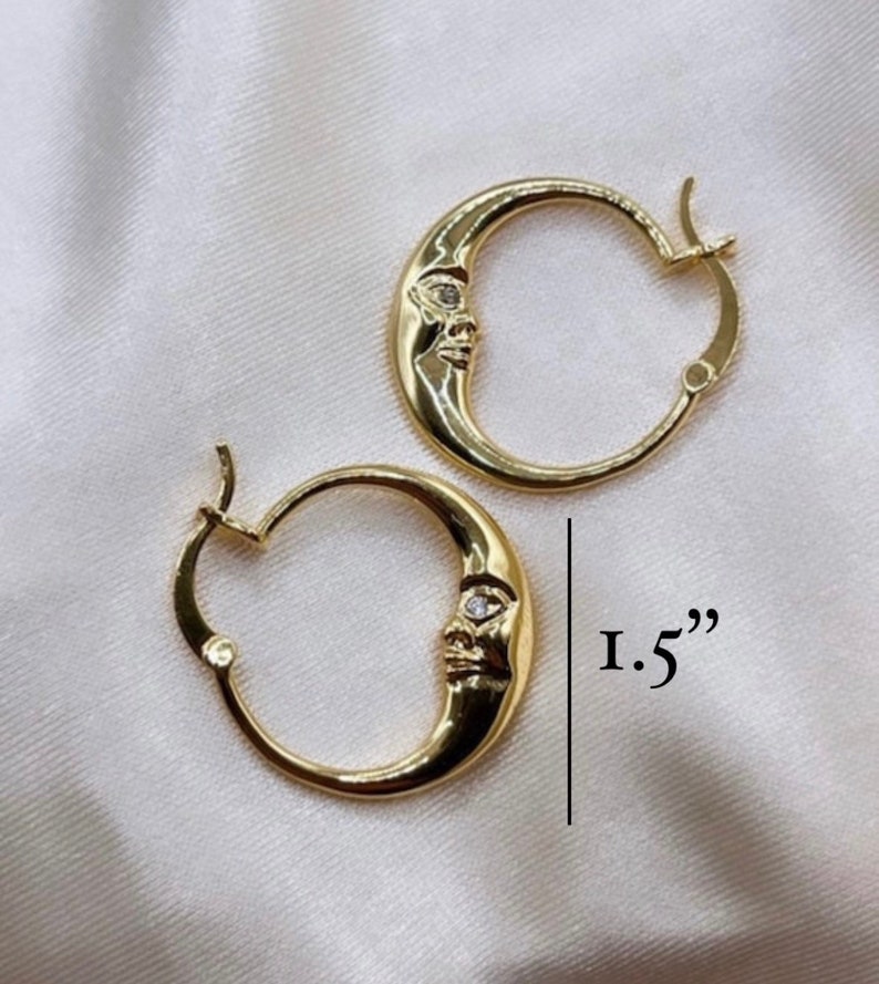 May include: A pair of gold-colored hoop earrings. Each earring features a crescent moon design with a face and a small, clear stone. The earrings measure 1.5 inches in length.