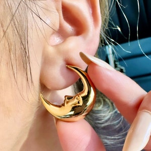 May include: Gold-coloured crescent moon earrings with a face design. The earrings are a shiny gold colour and are worn on the ear. The earrings are a unique and stylish accessory.