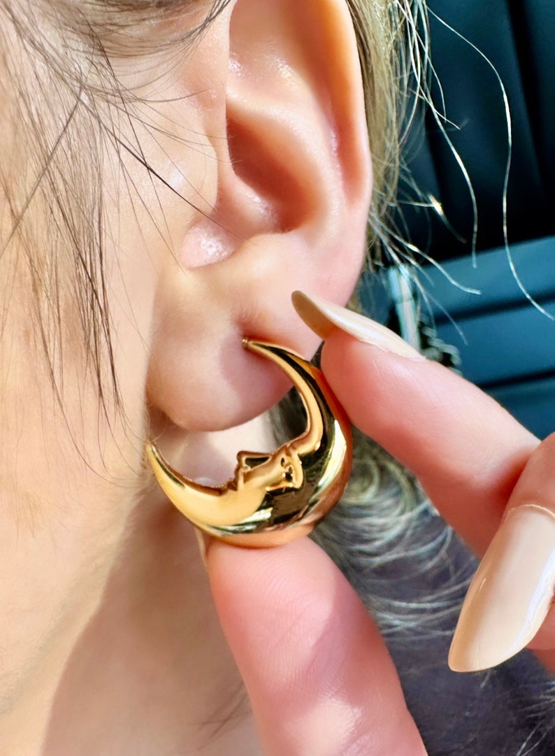 May include: Gold-coloured crescent moon earrings with a face design. The earrings are a shiny gold colour and are worn on the ear. The earrings are a unique and stylish accessory.