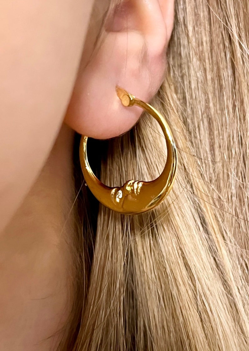 May include: Gold-colored crescent moon hoop earring. The earring features a detailed moon face design. The earring is worn on the ear. The background is out of focus.