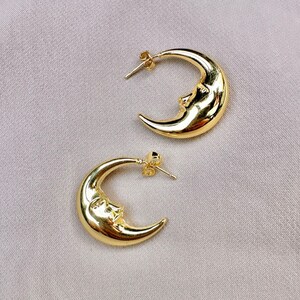 May include: A pair of gold-coloured crescent moon earrings. Each earring features a smiling face on the moon's surface. The earrings have a post-back closure and are displayed on a light grey fabric.