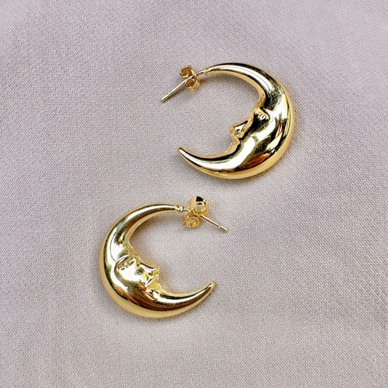 May include: A pair of gold-coloured crescent moon earrings. Each earring features a smiling face on the moon's surface. The earrings have a post-back closure and are displayed on a light grey fabric.