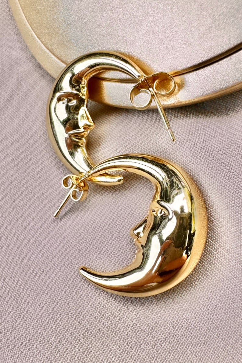 May include: A pair of gold-coloured crescent moon earrings. Each earring features a detailed face within the moon's curve. The earrings have a post-back closure and are displayed on a neutral background.