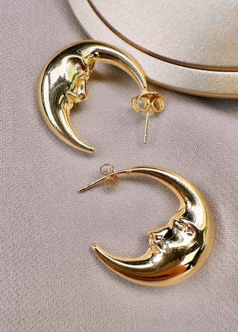 May include: A pair of gold-coloured crescent moon earrings. Each earring features a detailed face design on the moon's curve. The earrings have a post-back closure. The background is a neutral, textured surface.