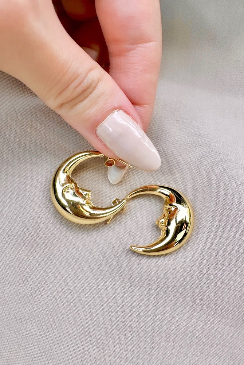May include: A pair of gold crescent moon earrings. Each earring has a detailed face within the moon's curve. The earrings are shown against a neutral background.