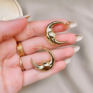 May include: A pair of gold-coloured crescent moon earrings. Each earring features a detailed face design on the moon's surface. The earrings have a post-back closure. The jewellery is displayed against a neutral background.