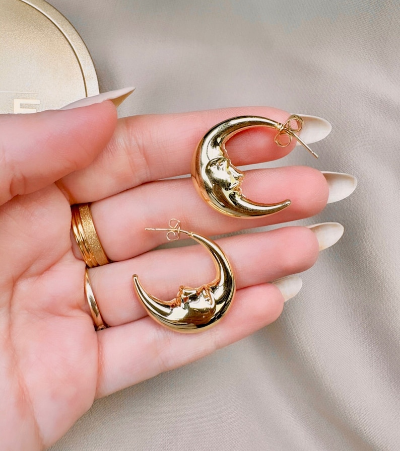 May include: A pair of gold-coloured crescent moon earrings. Each earring features a detailed face design on the moon's surface. The earrings have a post-back closure. The jewellery is displayed against a neutral background.