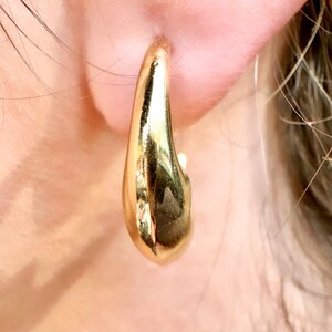 May include: A close-up of a gold-coloured hoop earring. The earring has a smooth, polished surface and a teardrop shape. The earring is worn on an ear, with the skin visible in the background.