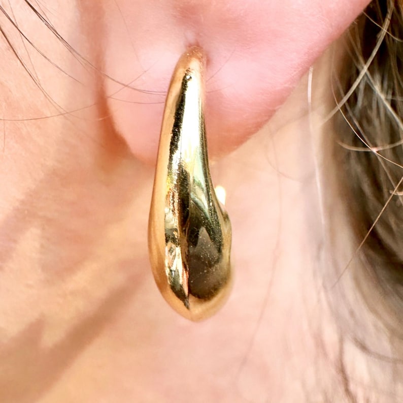 May include: A close-up of a gold-coloured hoop earring. The earring has a smooth, polished surface and a teardrop shape. The earring is worn on an ear, with the skin visible in the background.