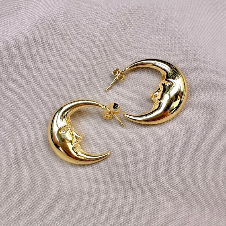 May include: A pair of gold-coloured crescent moon earrings. Each earring features a detailed face carved into the moon's surface. The earrings have a post-and-clutch closure. The background is a neutral, light tan colour.