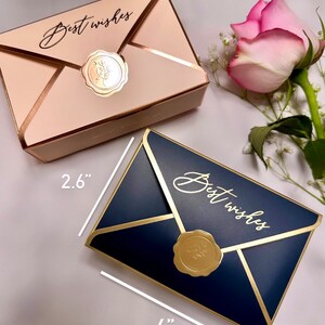 May include: Two envelope-shaped gift boxes, one beige and one navy blue, with gold accents and a gold seal that reads "Best wishes." The navy box measures 4 inches long. A pink rose and baby's breath are in the background.