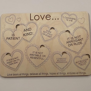 Puede incluir: Una placa de madera con un diseño en forma de corazón, que presenta la palabra "Love" y una lista de cualidades asociadas con el amor, incluyendo "paciente", "amable", "no envidia ni se jacta", "no es arrogante ni grosero", "no insiste en su propio camino", "no es irritable ni resentido", "no se regocija en la maldad", y "pero se regocija con la verdad". La placa también incluye el versículo bíblico 1 Corintios 13:4-8 y la frase "El amor todo lo sufre, todo lo cree, todo lo espera, todo lo soporta."