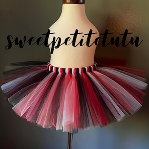 May include: A multi-layered tutu skirt in red, black, and white tulle. The waistband is striped with red, black, and white. The text "sweetpetitetutu" is written in cursive across the top of the skirt.