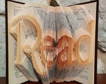 May include: A book art sculpture of the word "remarkable" created by folding the pages of a book. The sculpture is displayed on a wooden surface.