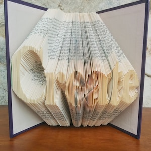 May include: A book art sculpture of the word "Orville" created by folding the pages of a book. The book is open and the pages are folded to create the word. The book has a purple cover.