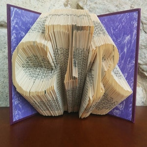 Könnte beinhalten: Ein Buch, das in die Form des Wortes "CIR" gefaltet ist, mit einem violetten Einband. Das Buch ist geöffnet und die Seiten sind gefaltet, um die Buchstaben zu bilden.