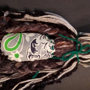 May include: A decorative tassel ornament featuring a central fabric piece with a green, white, and dark blue paisley and floral pattern. The ornament is surrounded by long, textured yarn strands in shades of brown, black, gray, and white, tied with a green cord.