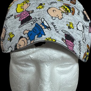 May include: A gray cap featuring a repeating pattern of Peanuts characters, including Snoopy, Charlie Brown, Lucy, and Sally. The cap has a curved brim and a gray trim. The characters are in various poses, with their names printed around them.