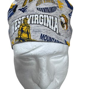 May include: A gray and blue West Virginia Mountaineers scrub cap. The cap features the words "West Virginia Mountaineers" in white and blue, along with various graphics and logos. The cap is made of a soft, patterned fabric.