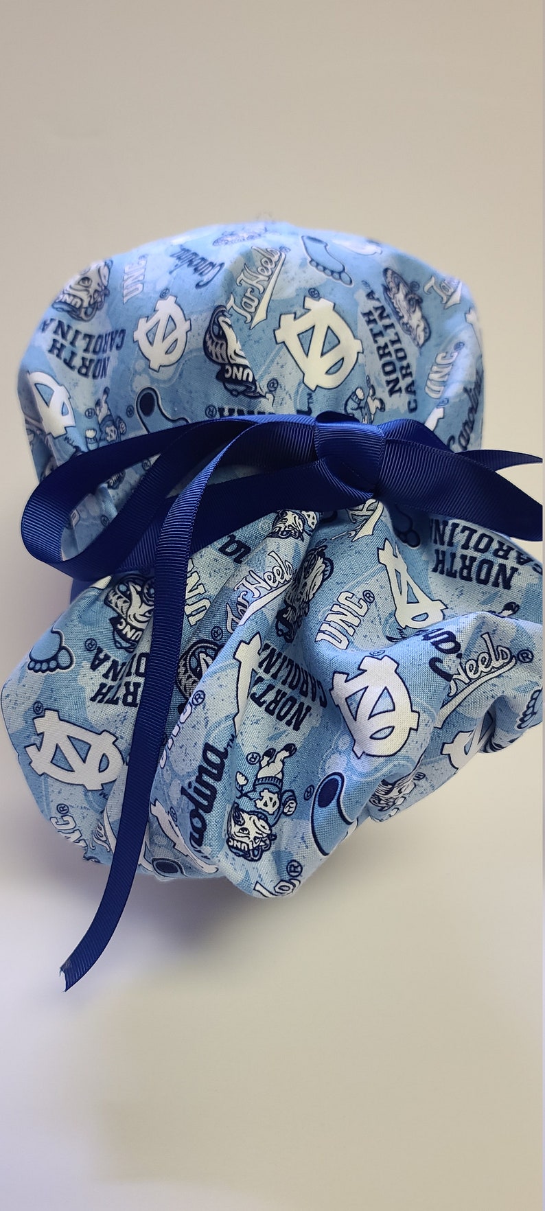 May include: A blue and white fabric scrub hat with a blue ribbon tied around it. The fabric features a repeated pattern of the University of North Carolina Tar Heels logo, the words "Tar Heels", "UNC", and "North Carolina".