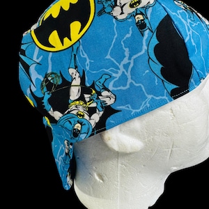 May include: A blue and black Batman-themed welding cap. The cap features a repeating pattern of the Batman logo and illustrations of Batman in action against a blue background with white lightning bolts. The cap is made of fabric.