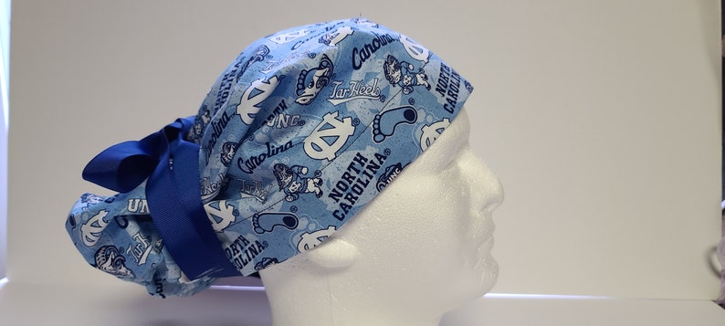 May include: Blue and white surgical scrub hat with a North Carolina Tar Heels pattern. The hat has a blue ribbon tied at the back.