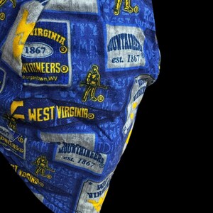 May include: Blue fabric with repeating patterns of West Virginia Mountaineers logos, text, and figures. The design includes the words "Mountaineers" and "West Virginia" in yellow and white, with the establishment year 1867.