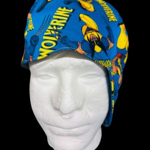 May include: A blue scrub cap featuring a repeating comic book print of the character Wolverine in a yellow and black suit, with the word Wolverine printed in bold yellow capital letters across the fabric, shown on a white mannequin head.