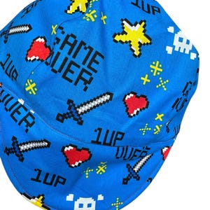 May include: A blue welding cap with a pixelated video game theme. The cap features the words "GAME OVER" and "1UP", along with pixelated hearts, swords, stars, and skull graphics. The cap is designed with a curved brim and a red trim.