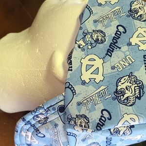 May include: A blue and white patterned welding cap with the text "North Carolina" and "UNC" repeated throughout the design. The cap has a flap that covers the ears and neck.