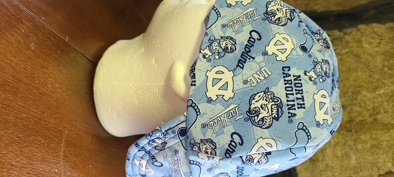 May include: A blue and white patterned welding cap with the text "North Carolina" and "UNC" repeated throughout the design. The cap has a flap that covers the ears and neck.