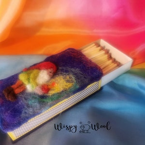 Könnte beinhalten: Ein gefilzter Zwerg mit roter Mütze und grüner Jacke liegt auf einer blauen Oberfläche neben einer Streichholzschachtel. Der Hintergrund ist ein weicher Farbmix. Der Text "Wispy Wool" ist im Bild enthalten.