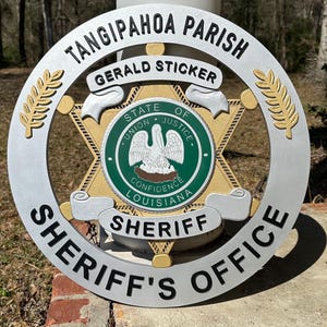 May include: A silver and gold Sheriff's Office emblem with the text "TANGIPAHOA PARISH" and "SHERIFF'S OFFICE". The emblem features a star shape with a green and gold center seal that reads "STATE OF LOUISIANA".