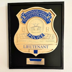 May include: A framed wooden plaque featuring a police badge design. The badge is tan with blue lettering that reads "PENTAGON FORCE PROTECTION AGENCY POLICE LIEUTENANT." The plaque is set against a black background.